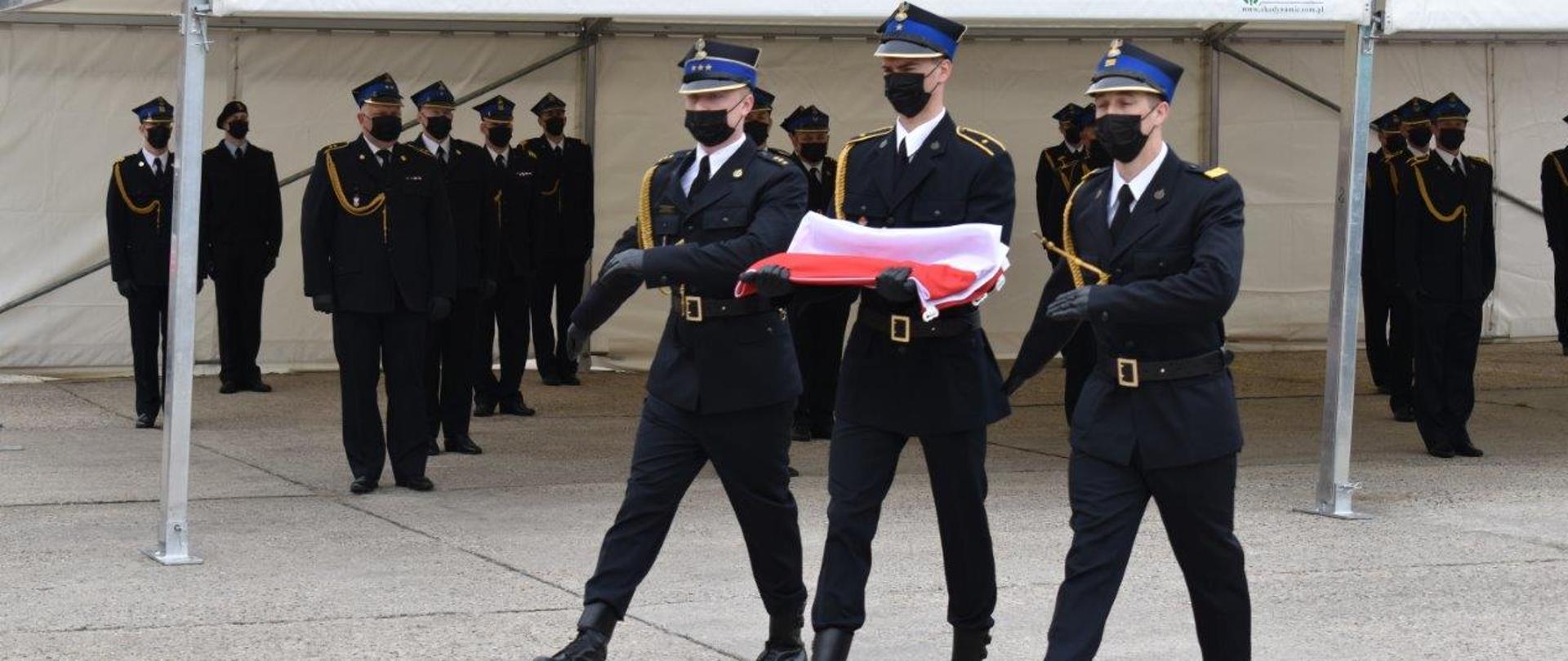 Poczet flagowy maszeruje w kierunku masztu. W tle funkcjonariusze KP PSP w Zgorzelcu wskazania do odznaczeń oraz awansów z okazji Dnia Strażaka. Funkcjonariusze w umundurowaniu wyjściowym.