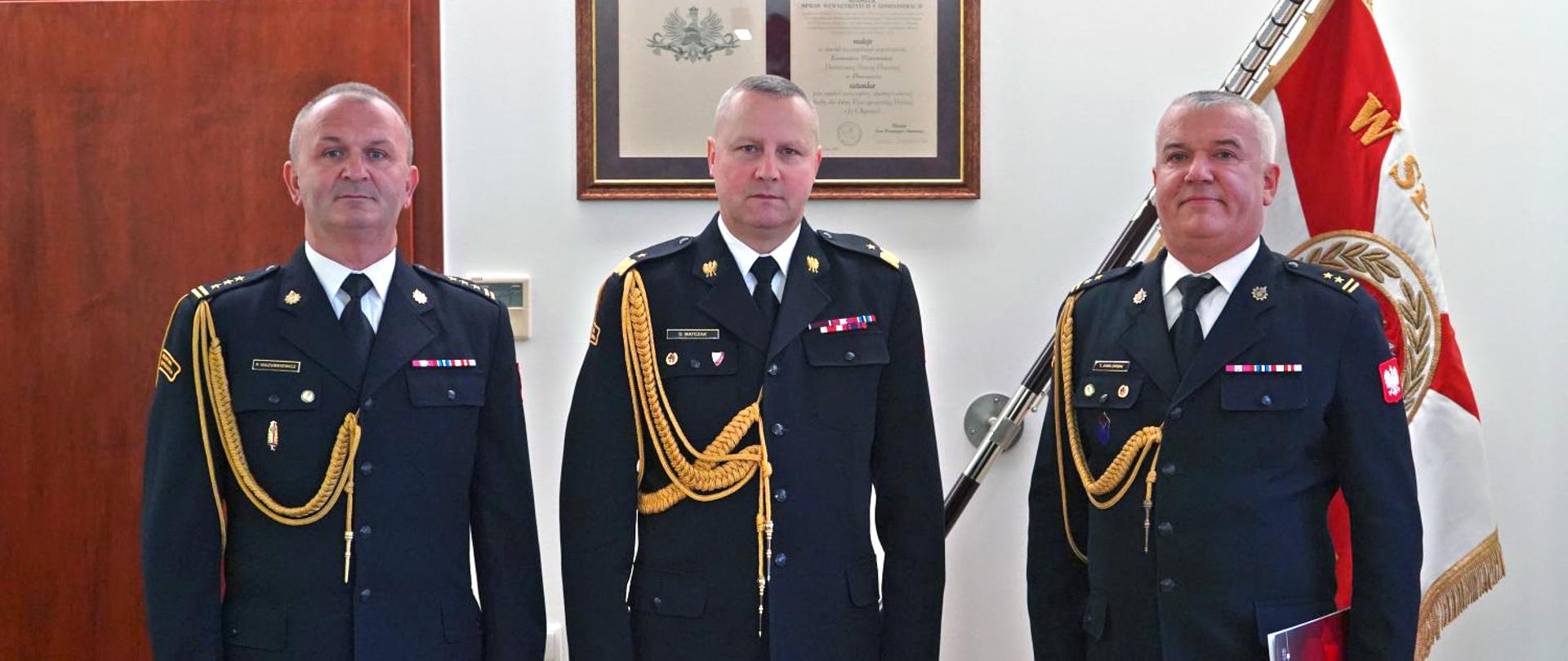 Strażacy w mundurach wyjściowych w trakcie uroczystego spotkania. W tle sztandar komendy.