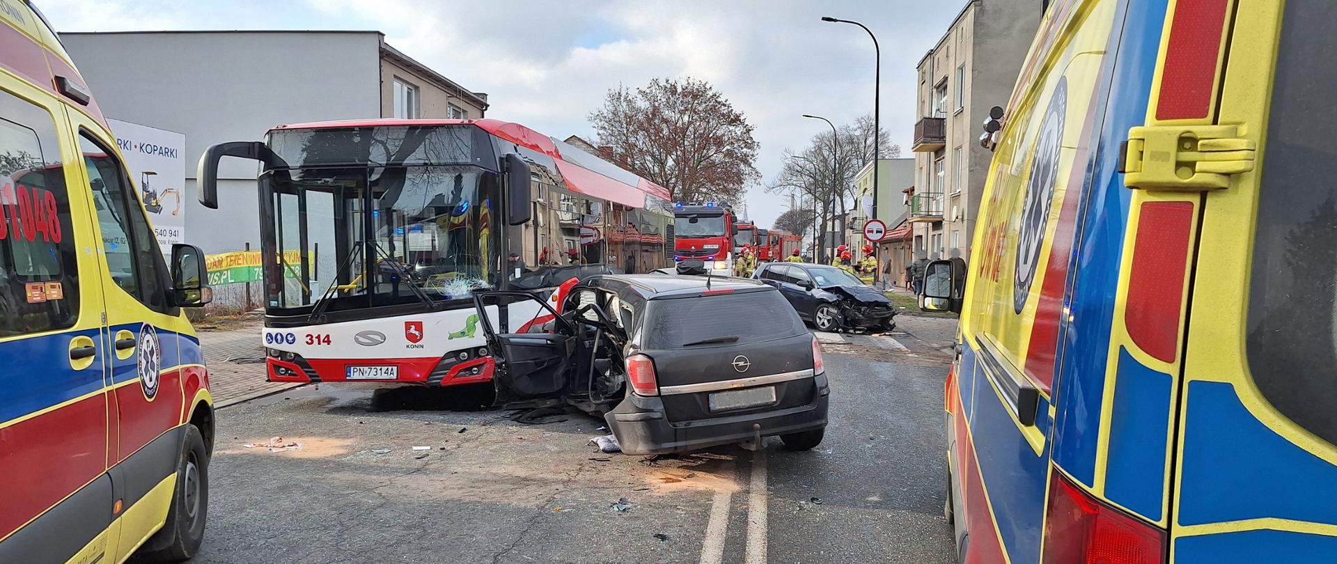 Samochód osobowy marki OPEL stoi na środku jezdni i przy autobusie MZK. Z tyłu widać drugi samochód osobowy marki VW z uszkodzonym przodem. Za pojazdami osobowymi stoją samochody strażackie. Z przodu widać karetki pogotowia ratunkowego, 