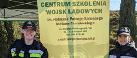 dwie kobiety strażak ubrane w czarne mundury i czapeczki typu dżokejka stoją przy banerze wojskowym