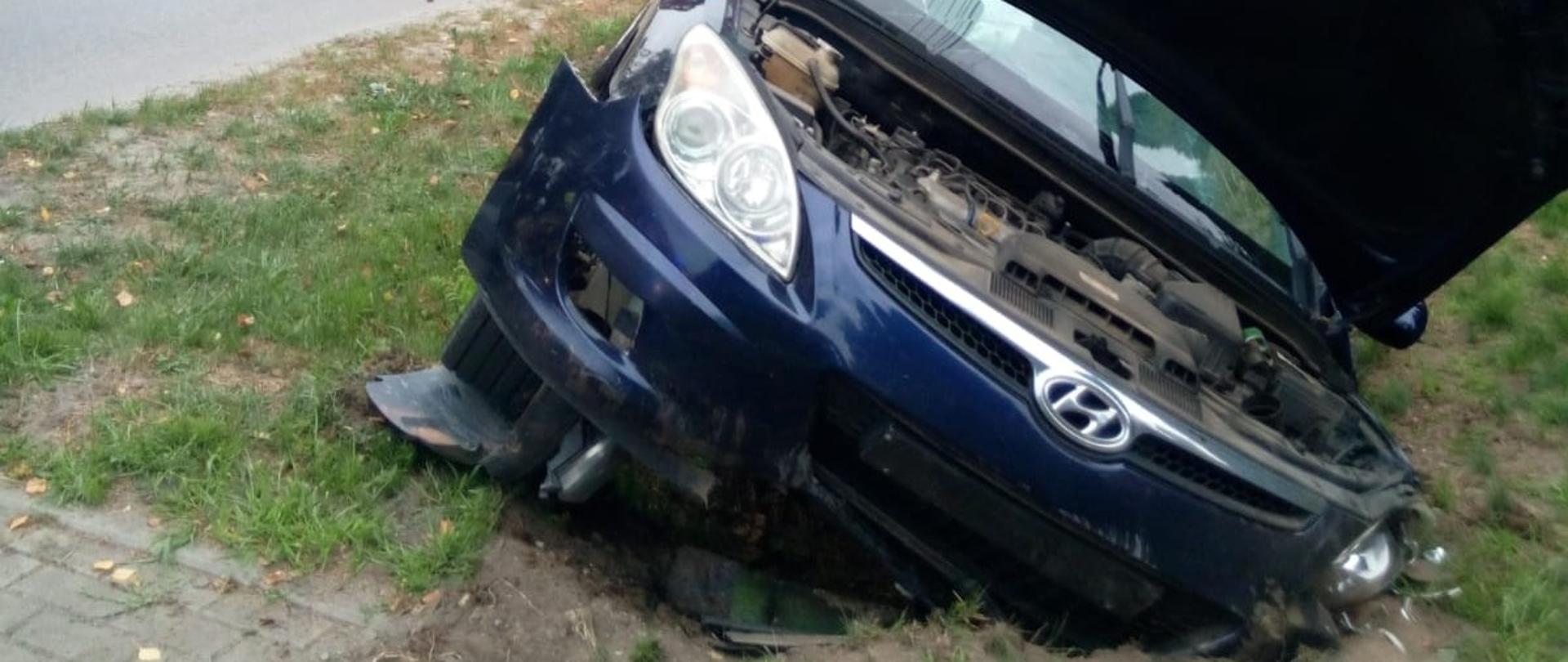 Zdjęcie przedstawia samochód osobowy znajdujący się w przydrożnym rowie. Za nim stoi samochód pożarniczy i ratownicy.