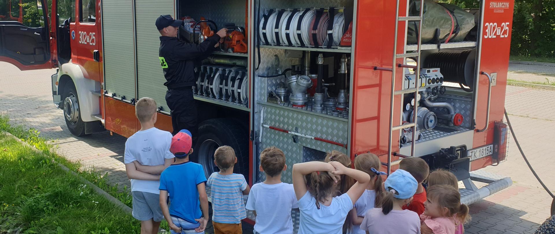 Zdjęcie przedstawia samochód pożarniczy podczas wizyty na wakacyjnej półkolonii. Przy aucie stoi strażak a wokół stoją słuchające strażaka dzieci.