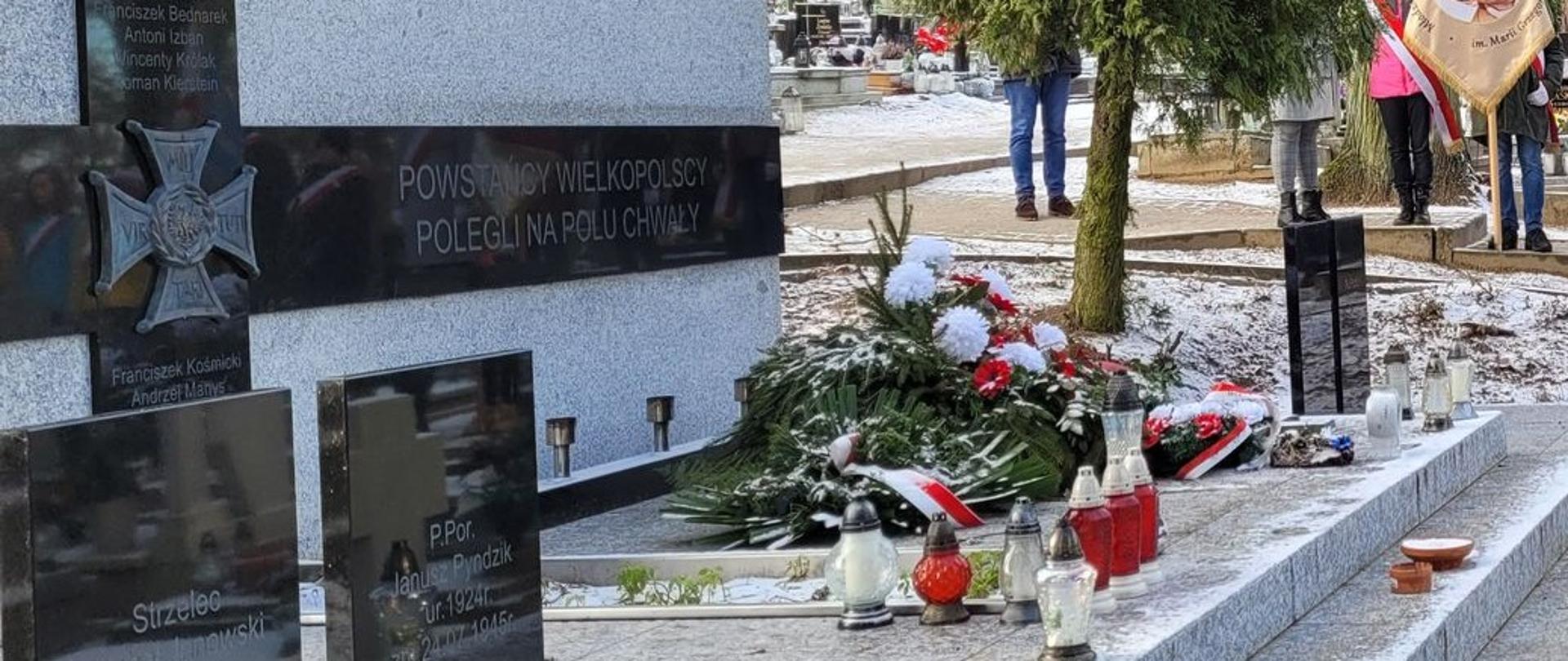 Zdjęcie przedstawia delegacje strażaków podczas uroczystego capstrzyka przy powstańczej mogile.
W tle drzewa i groby na cmentarzu.
