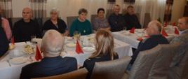 Świąteczne spotkanie strażackich emerytów. Sala w jednej z rawickich restauracji. Po obu stronach stołu zasiadają uczestnicy spotkania, mężczyźni i kobiety. Na stole są białe filiżanki, szklanki, talerzyki, sztućce i dzbanki oraz czerwone serwetki i surówki.