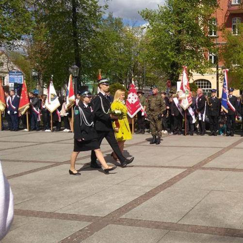 Zdjęcie przedstawia dziewczynę i chłopaka w mundurach Ochotniczej Straży Pożarnej oraz nauczycielkę w żółtym płaszczu. Trójka osób składa kwiaty pod pomnikiem poświęconym Poległym za Wolną i Niezawisłą Polskę. W tle delegacje grup ze sztandarami.