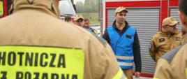 Na zdjęciu widoczny jest funkcjonariusz PSP ubrany w niebieską kamizelkę na tle samochodu strażackiego podczas odprawy ze strażakami ochotnikami przed rozpoczęciem ćwiczeń.