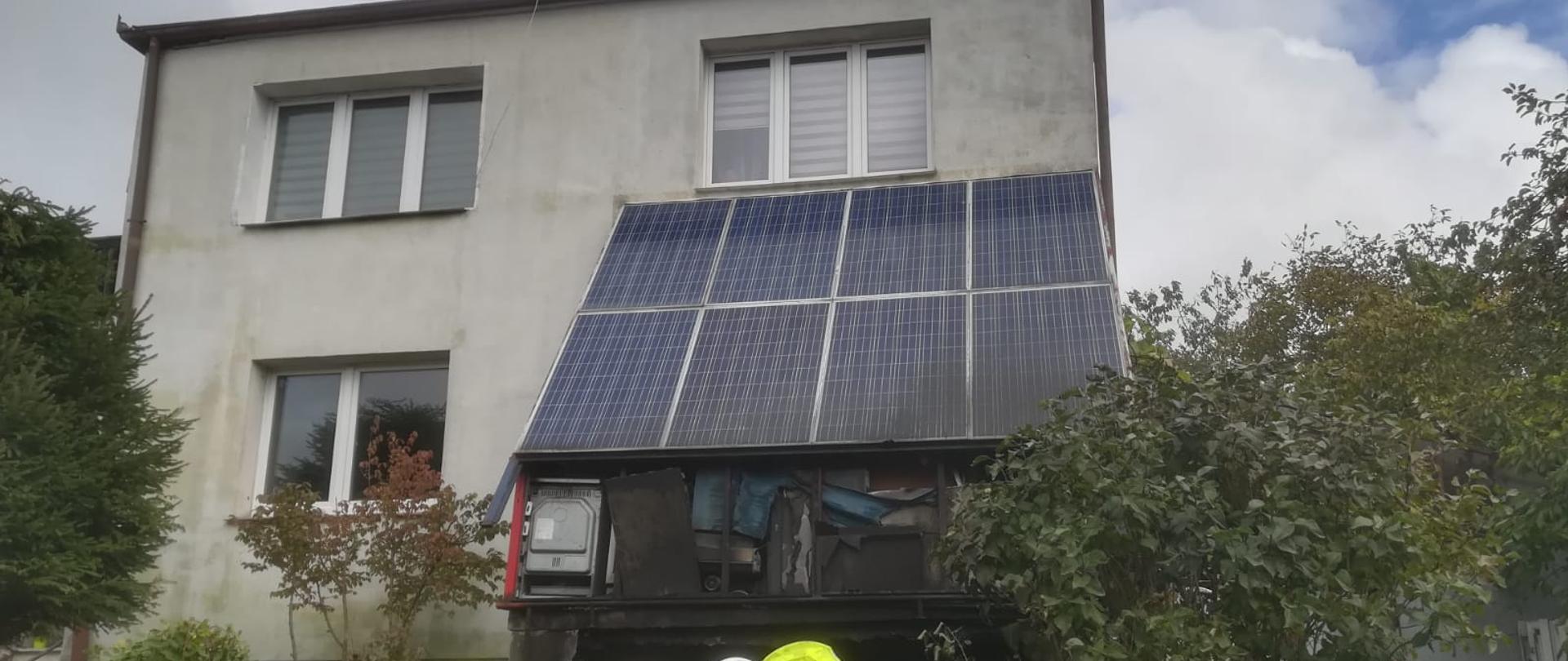 Budynek mieszkalny po pożarze. Na pierwszym planie 4 strażaków ubranych w ubrania specjalne podczas działań ratowniczo gaśniczych. W tle zdjęcia widok budynku mieszkalnego, z bryły budynku wystaje przybudówka z dachem zrobionym z paneli fotowoltaicznych. Z przybudówki wystają spalone przedmioty (sprzęt AGD i RTV) 
