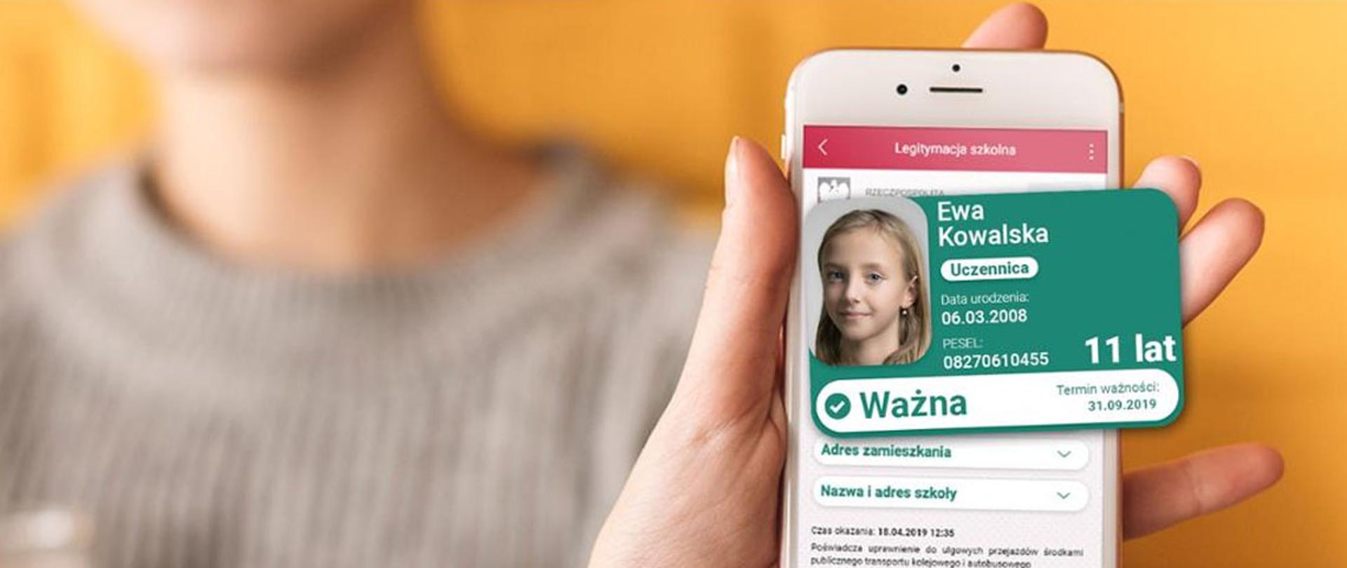 Osoba trzymająca w wyciągniętej ręce telefon. Na pierwszym planie, po prawej stronie dłoń z ekranem na którym wyświetlona jest aplikacja mlegitymacja z fikcyjnymi danymi