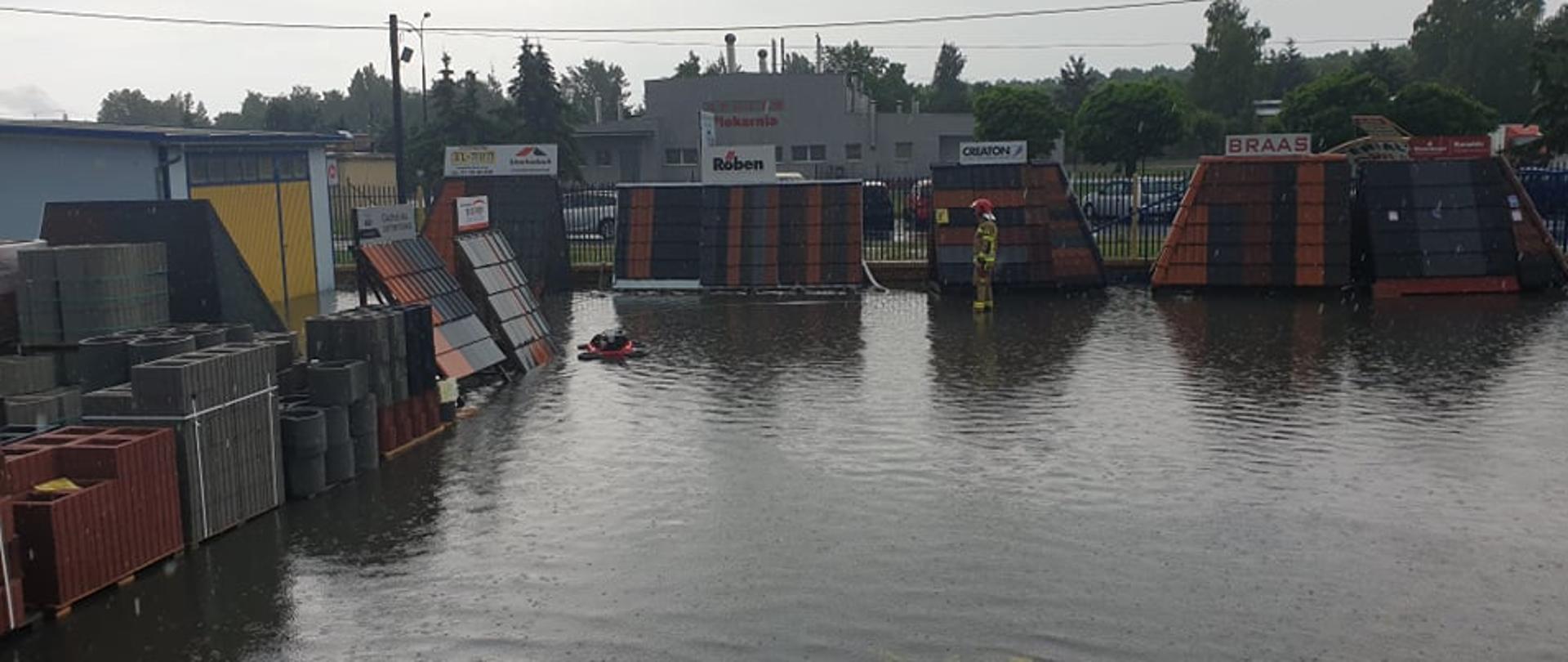 Na zdjęciu zalany plac budowlany przez ulewne deszcze, pompa pracująca celem wypompowania wody oraz strażak wykonujący czynności ratownicze 