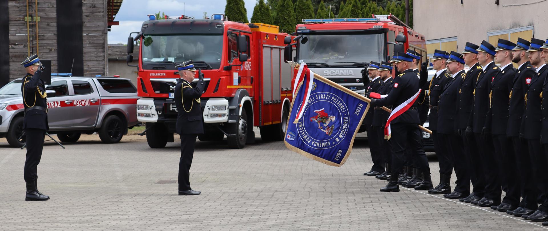 Strażacy w galowych mundurach stoją w szyku podczas uroczystego apelu. Na pierwszym planie poczet sztandarowy prezentuje sztandar jednostki, a przed nim strażak oddaje honor. W tle widoczne są czerwone samochody pożarnicze zaparkowane na placu przed remizą.