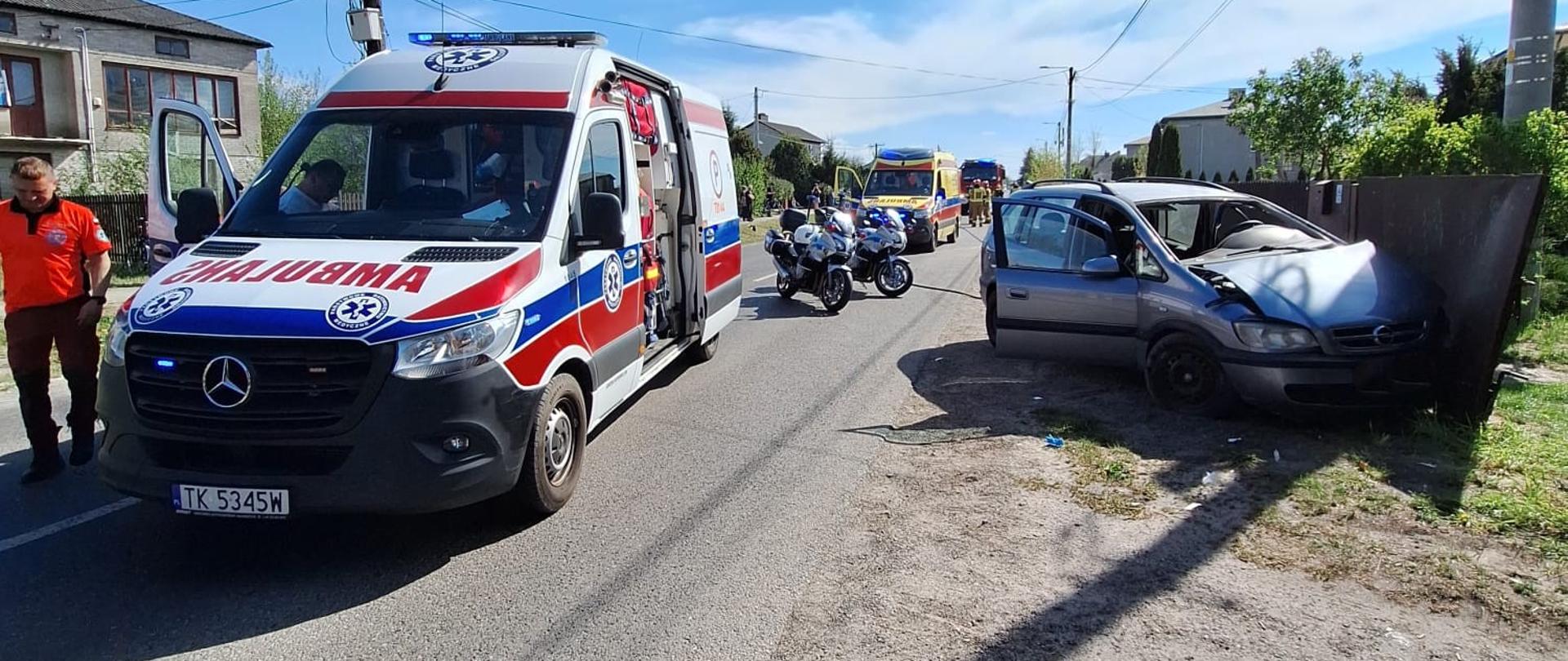 Zdjęcie przedstawia wypadek drogowy z udziałem szarego samochodu osobowego marki Opel, który uderzył przodem w metalowe ogrodzenie i zatrzymał się na trawiastym poboczu. Przednie drzwi od strony kierowcy są otwarte.