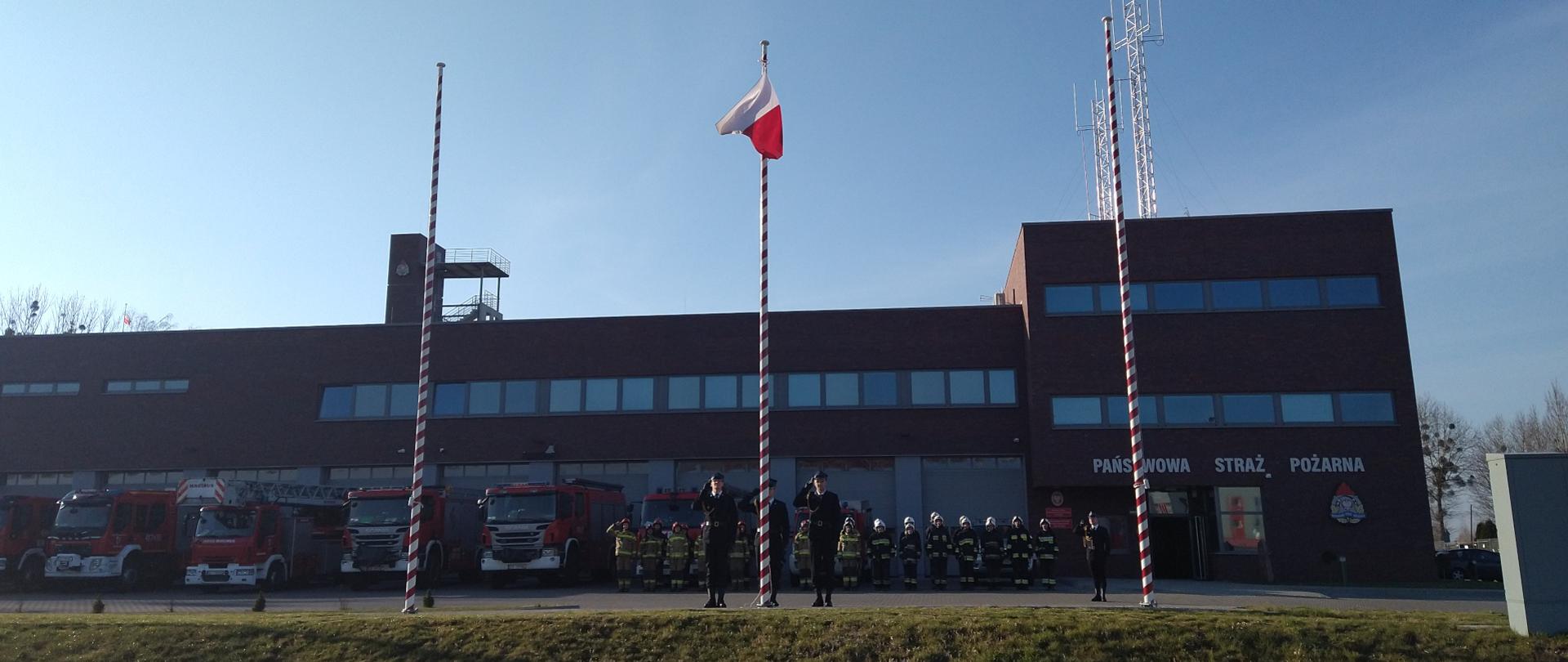 Zdjęcie przedstawia poczet flagowy podnoszący flagę państwową na maszt. Na drugim planie, na tle budynku komendy i samochodów pożarniczych stoją w szeregu strażacy w ubraniach specjalnych.