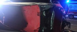 Zdjęcie obrazuje przewrócony na prawy bok samochód Ford Transit. Pojazd ma uszkodzony przód i rozbitą przednią szybę. W tle widać karetkę powiatową z włączonymi sygnałami świetlnymi.