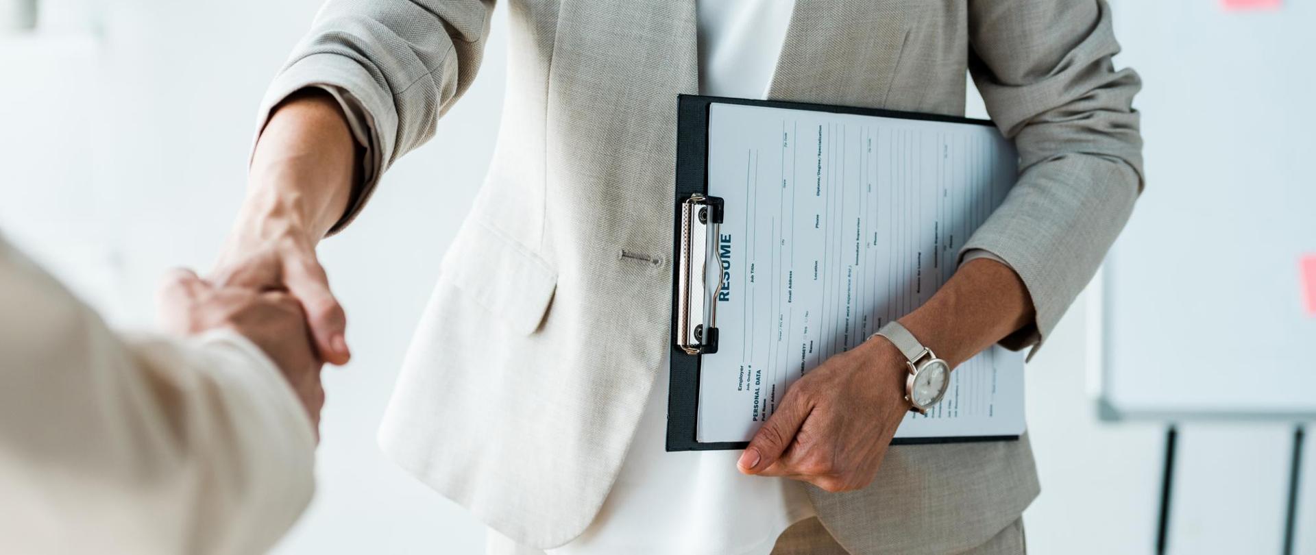 Na zdjęciu jest kobieta z teczką podająca rękę innej osobie. W tle jest tablica.