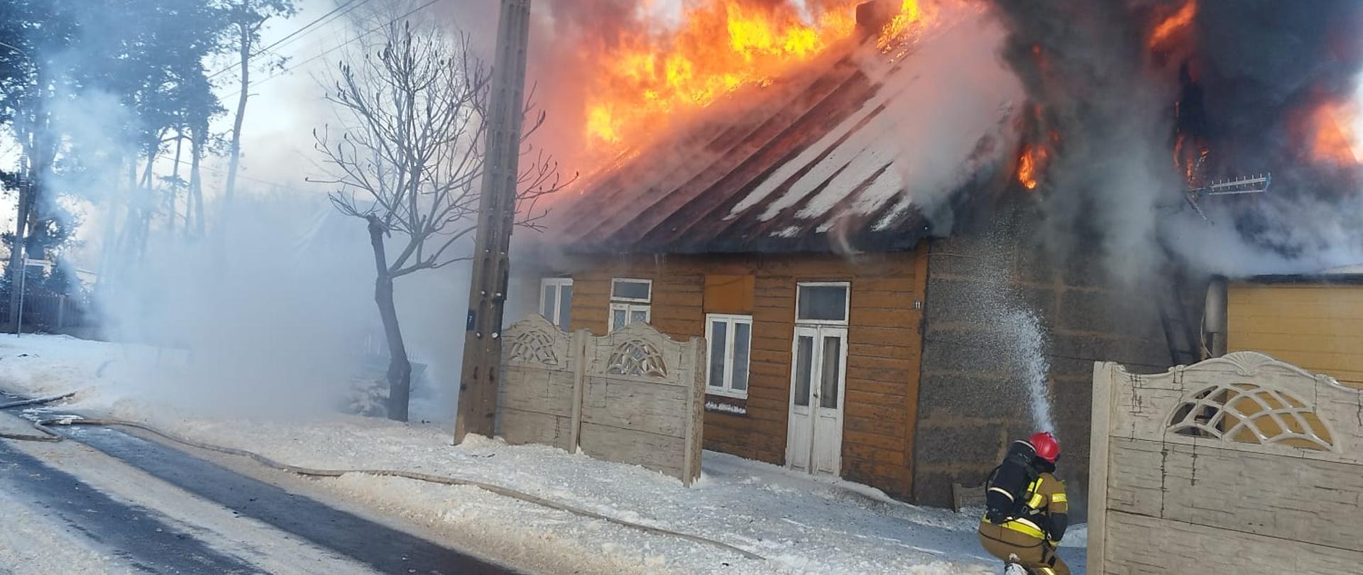 Pożar drewnianego budynku mieszkalnego. Jeden ze strażaków podaje prąd wody na palący się budynek, ma założony aparat powietrzny na plecach. Dookoła panuje zimowa aura oraz leży śnieg.