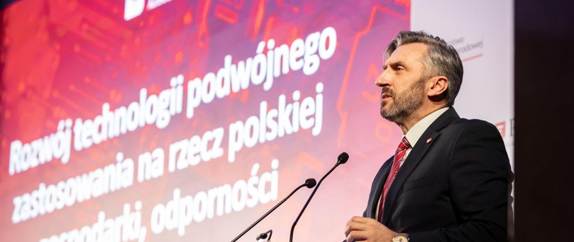 Wiceminister Waldemar Sługocki przemawia podczas IDA BootCamp. Wiceminister stoi przy mównicy, w tle prezentacja wydarzenia. 