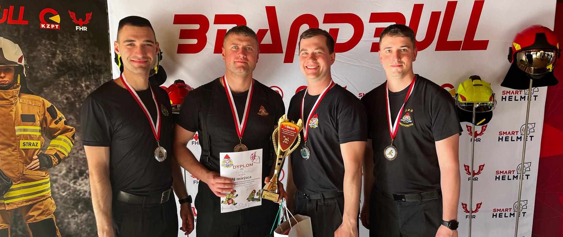 Zdjęcie przedstawia 4 osobowy zespół strażaków z medalami na szyi, pucharem i dyplomem oraz nagrodami rzeczowymi za zajęcie 2 miejsca w mistrzostwach wielkopolski w ratownictwie w ramach KPP