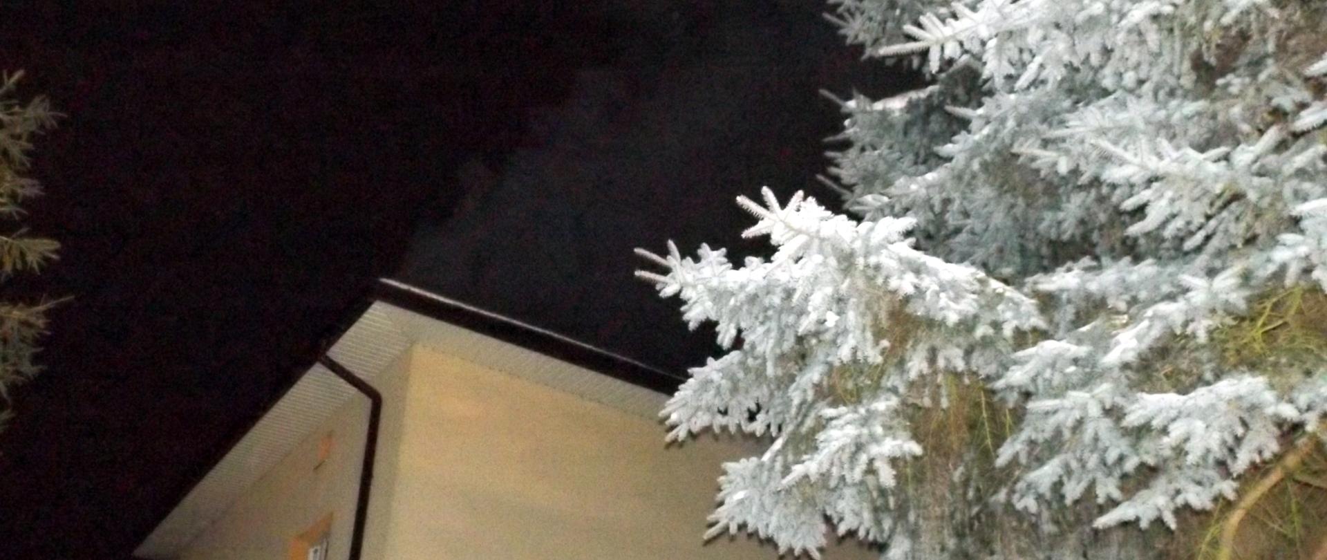 Zdjęcie przedstawia budynek mieszkalny - wysoki dom, obok znajduje się zaśnieżone drzewo. Z komina na tle nocnego nieba widać wydobywający się dym - prawdopodobnie z komina.