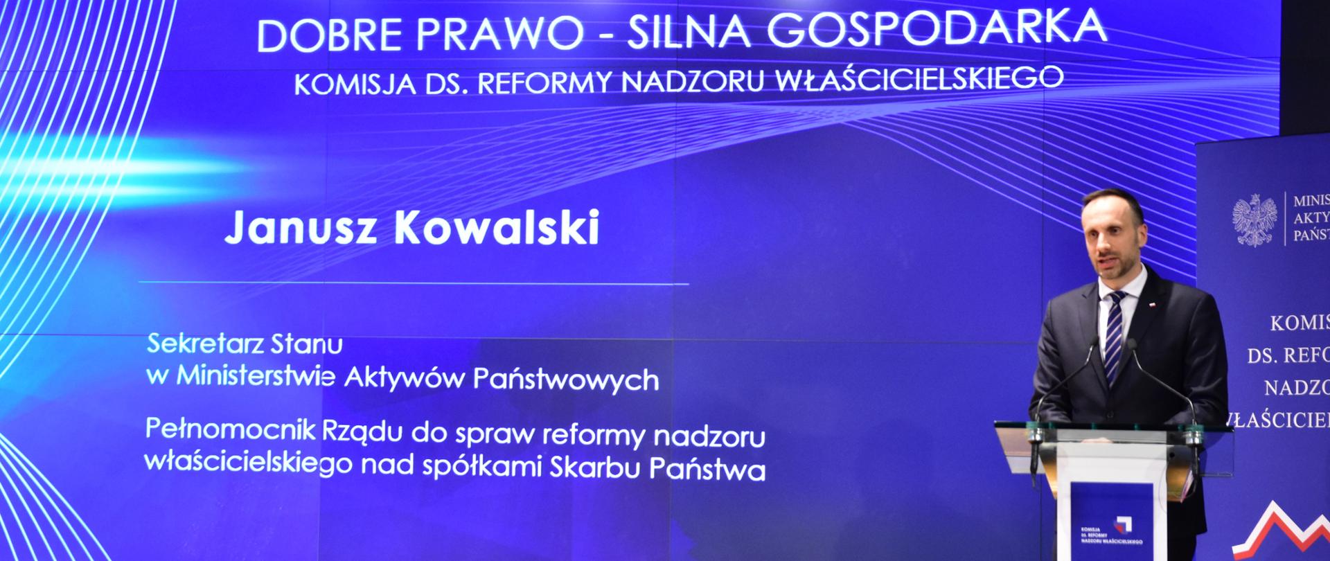 Wiceminister aktywów państwowych Janusz Kowalski przy mównicy. Z tyłu plansza z napisem Dobre prawo - silna gospodarka, komisja ds. reformy nadzoru właścicielskiego, imię i nazwisko ministra oraz funkcje pełnione w resorcie. U góry logotypy MAP i komisji ds. reformy nadzoru właścicielskiego