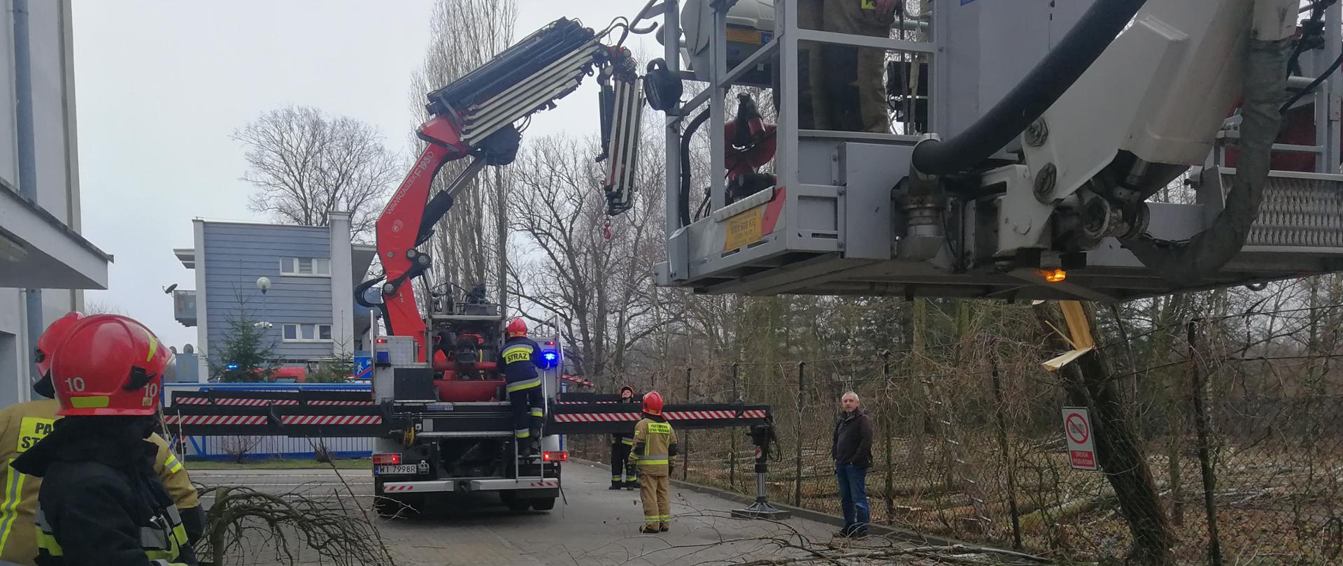 Na terenie działań widzimy kończących akcję strażaków. Kosz podnośnika opuszczany jest już na ziemię, a ramię dźwigu składane. Na ziemi widać pocięte i połamane kawałki drzewa które oparte było o budynek