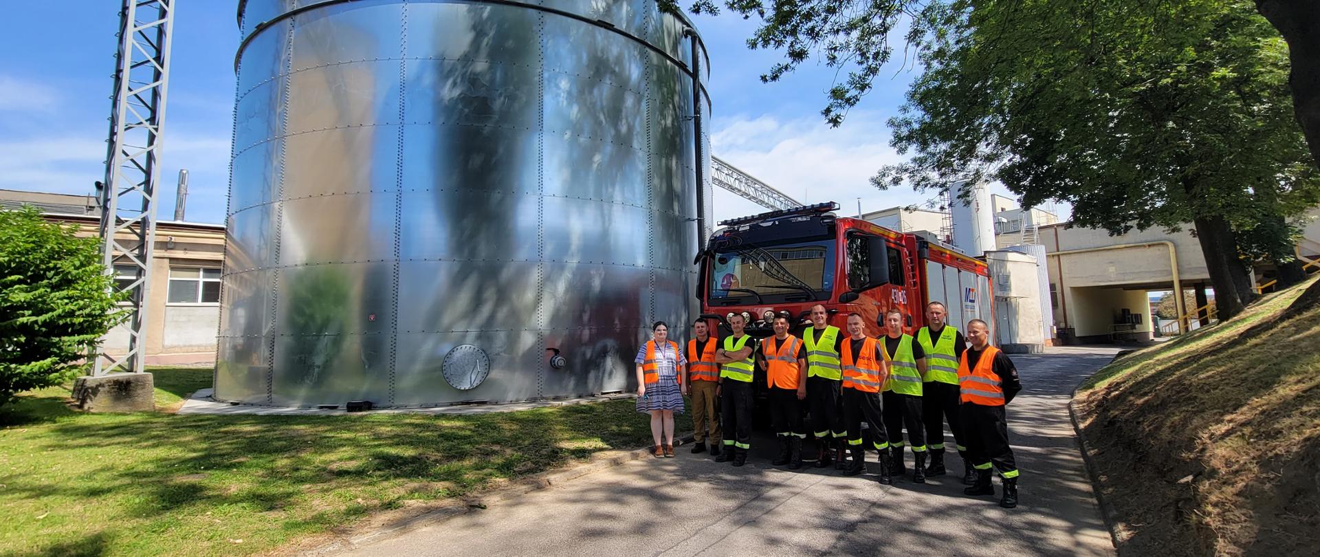Na zdjęciu widoczni są strażacy oraz kobieta z obsługi fabryki ustawieni do zdjęcia grupowego w dwuszeregu. Za nimi znajduje się wóz strażacki, a obok duży, cylindryczny zbiornik. Strażacy ubrani są w ubrania koszarowe oraz kamizelki odblaskowe.