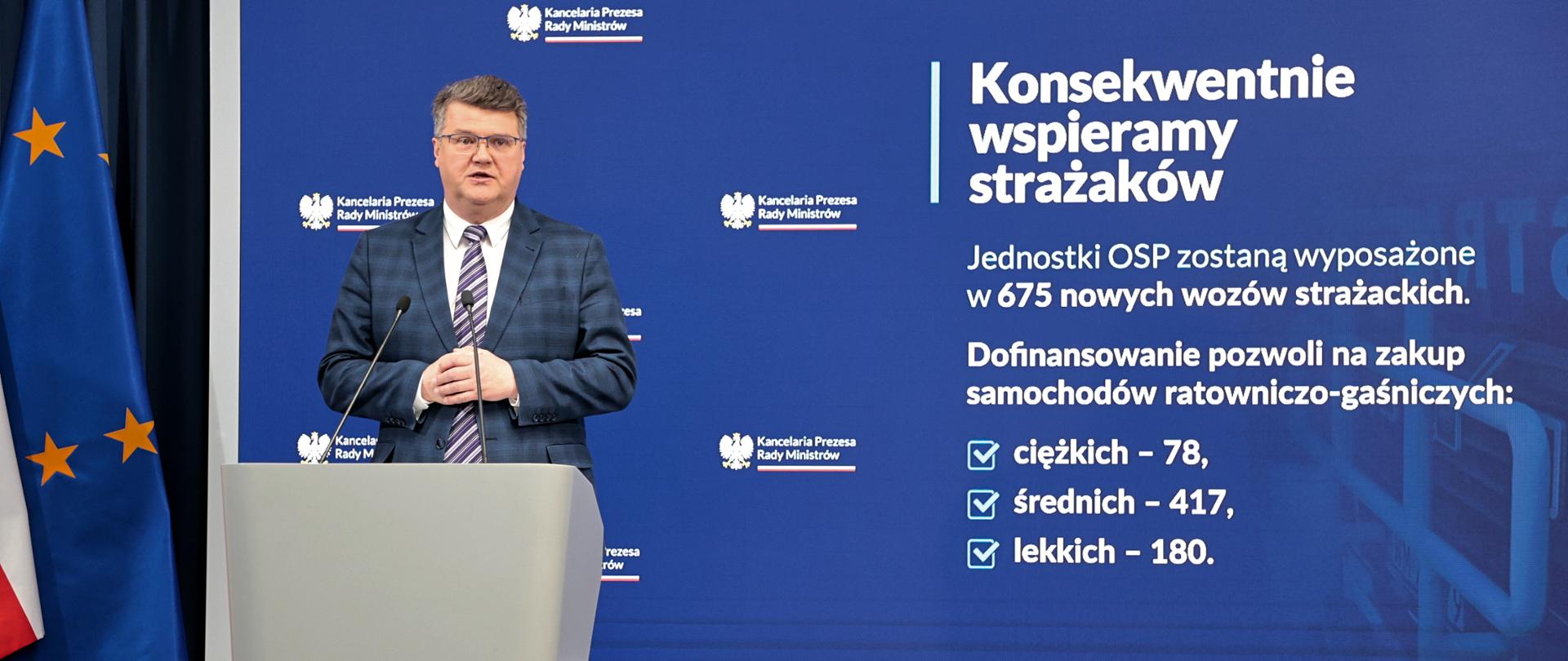 Maciej Wąsik przemawia przy mównicy w trakcie konferencji prasowej za nim banner mswia i napisami konsekwentnie wspieramy strażaków. Jednostki OSP zostaną wyposażone w 675 nowych wozów strażackich. dofinansowanie pozwoli na zakup samochodów ratowniczo-gaśniczych ciężkich 78 średnich 417 lekkich 180