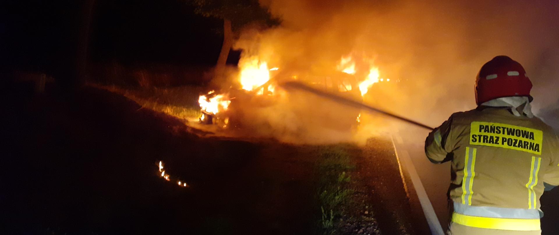 Na pierwszym planie widać strażaka PSP podającego prąd gaśniczy wody na płonący pojazd.