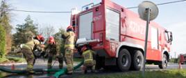 Widok z boku. Centralnie samochód pożarniczy. Z lewej strony czterech strażaków za samochodem sprawia linię ssawną. 