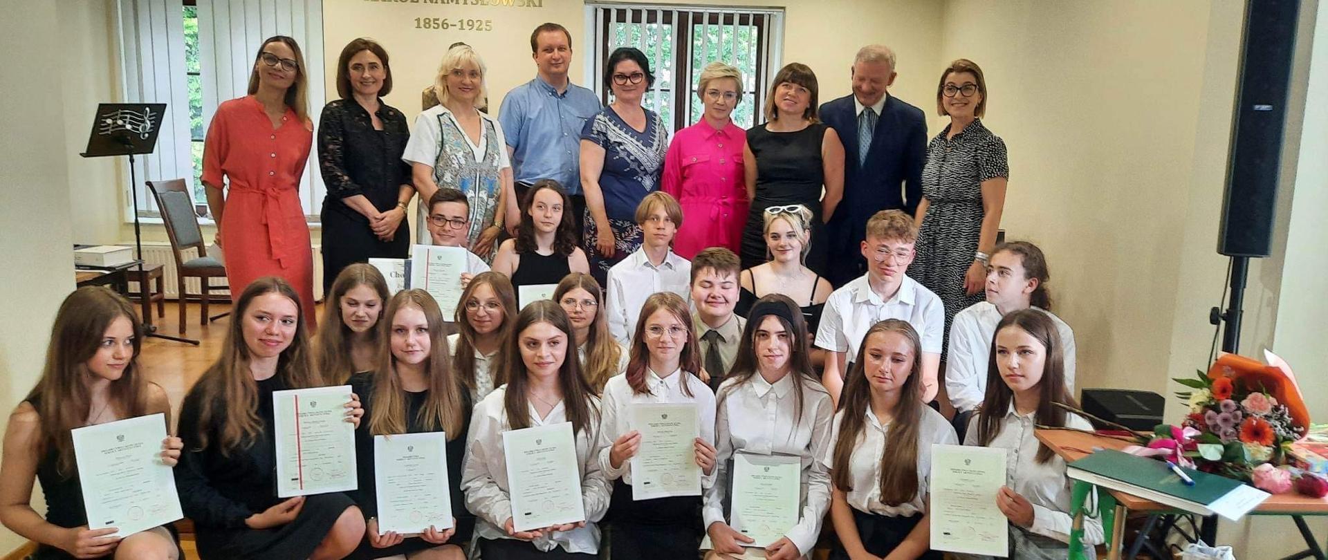 W sali koncertowej szkoły uczniowie klas kończących VI/6 i IV/4 po wręczeni świadectw ukończenia szkoły w dniu 22 czerwca 2023 r. Na scenie stoją nauczyciele, po prawej stronie pani dyr. Marta ostrowska. Niżej kucają w dwóch rzędach uczniowie ubrani na galowo, trzymają w rękach świadectwa.