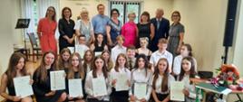 W sali koncertowej szkoły uczniowie klas kończących VI/6 i IV/4 po wręczeni świadectw ukończenia szkoły w dniu 22 czerwca 2023 r. Na scenie stoją nauczyciele, po prawej stronie pani dyr. Marta ostrowska. Niżej kucają w dwóch rzędach uczniowie ubrani na galowo, trzymają w rękach świadectwa.