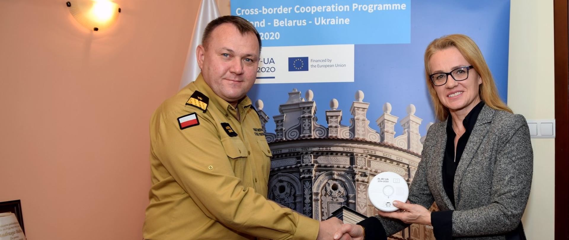 Zdjęcie zrobione w pomieszczeniu. Oficer Państwowej Straży Pożarnej w stopniu generała, stojący po lewej stronie zdjęcia, podaje rękę kobiecie trzymającej czujkę tlenku węgla i dymu. Za nimi stoi roll-up z nazwą projektu unijnego.