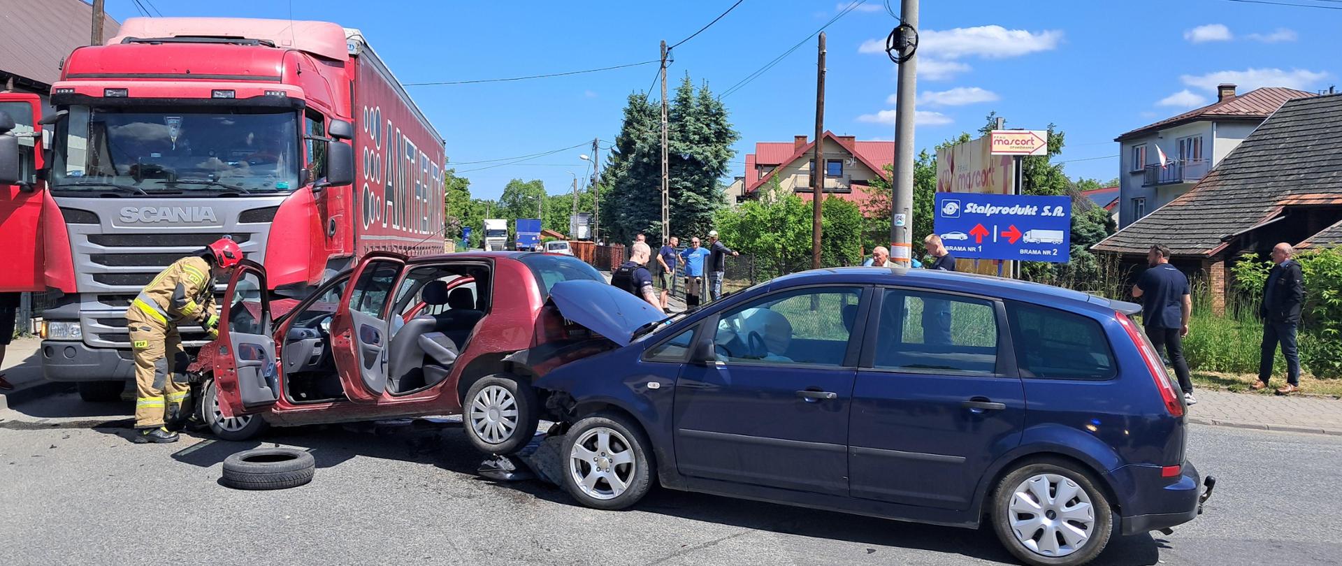 Zdjęcie przedstawia zderzenie trzech pojazdów – dwóch osobowych i jednego ciężarowego. Na zdjęciu widać przód czerwonego pojazd ciężarowego .Po prawej stronie widać czerwony samochód osobowy który uderzył przodem w samochód ciężarowy a w jego tył wjechał drugi niebieski samochód osobowy. Czerwony pojazd ma zniszczony przód, otwarte drzwi przednie i tylne oraz podniesiony tył, koła unoszą się nad drogą. Na zdjęciu widać drugi niebieski pojazd który ma uszkodzony przód i podniesiona maskę przednią. Po lewej stronie widać strażaka w piaskowym mundurze i czerwonym hełmie który odpina akumulator w pojeździe. Przed pojazdami leży opona. Z tył pojazdów widać osoby postronne.