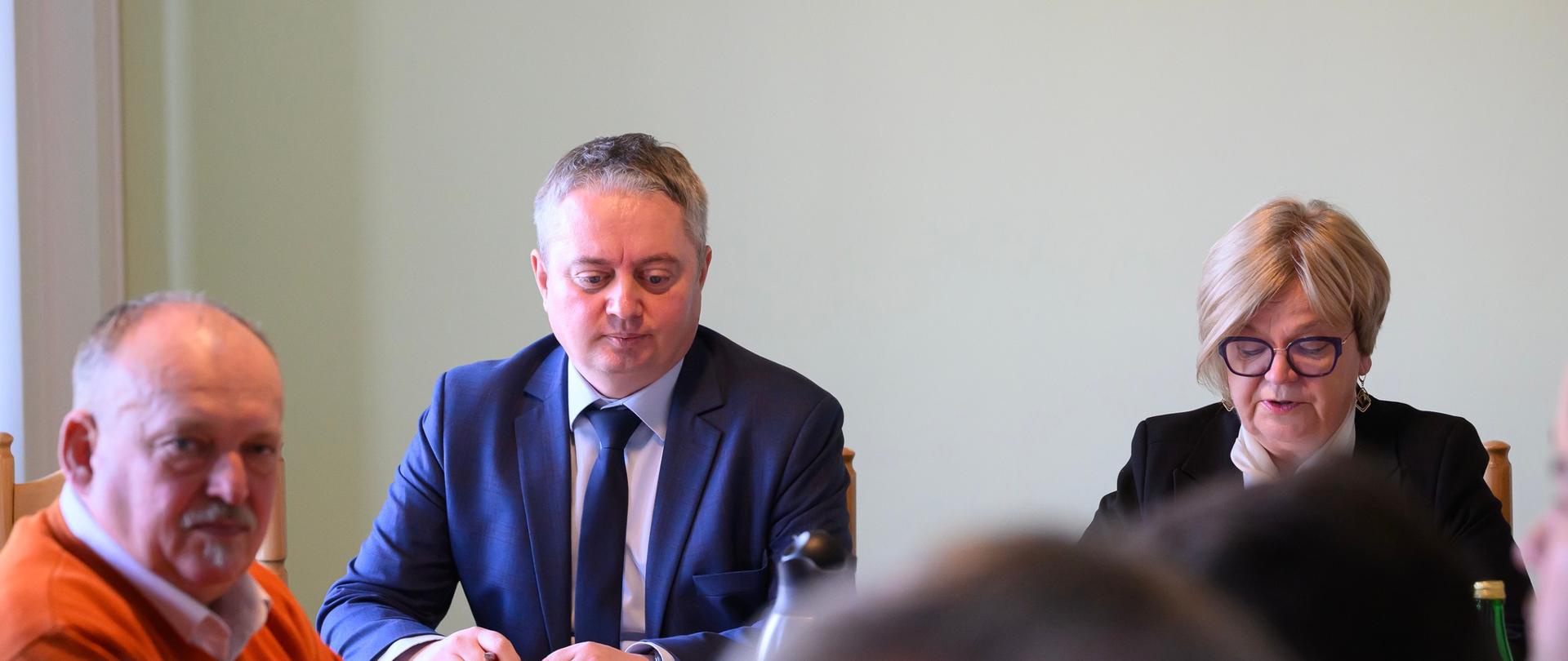 Siedzące przy stole trzy osoby. Dwóch mężczyzn po lewej stronie i kobieta po prawej stronie. Mężczyźni słuchają kobieta czyta.