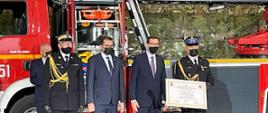 (od lewej) zastępca KG PSP, Wojewoda Małopolski, Premier RP oraz Komendant Powiatowy ustawieni w szeregu pozują do zdjęcia, komendant powiatowy trzyma w rękach akt przekazania, za nimi samochód ratowniczy PSP