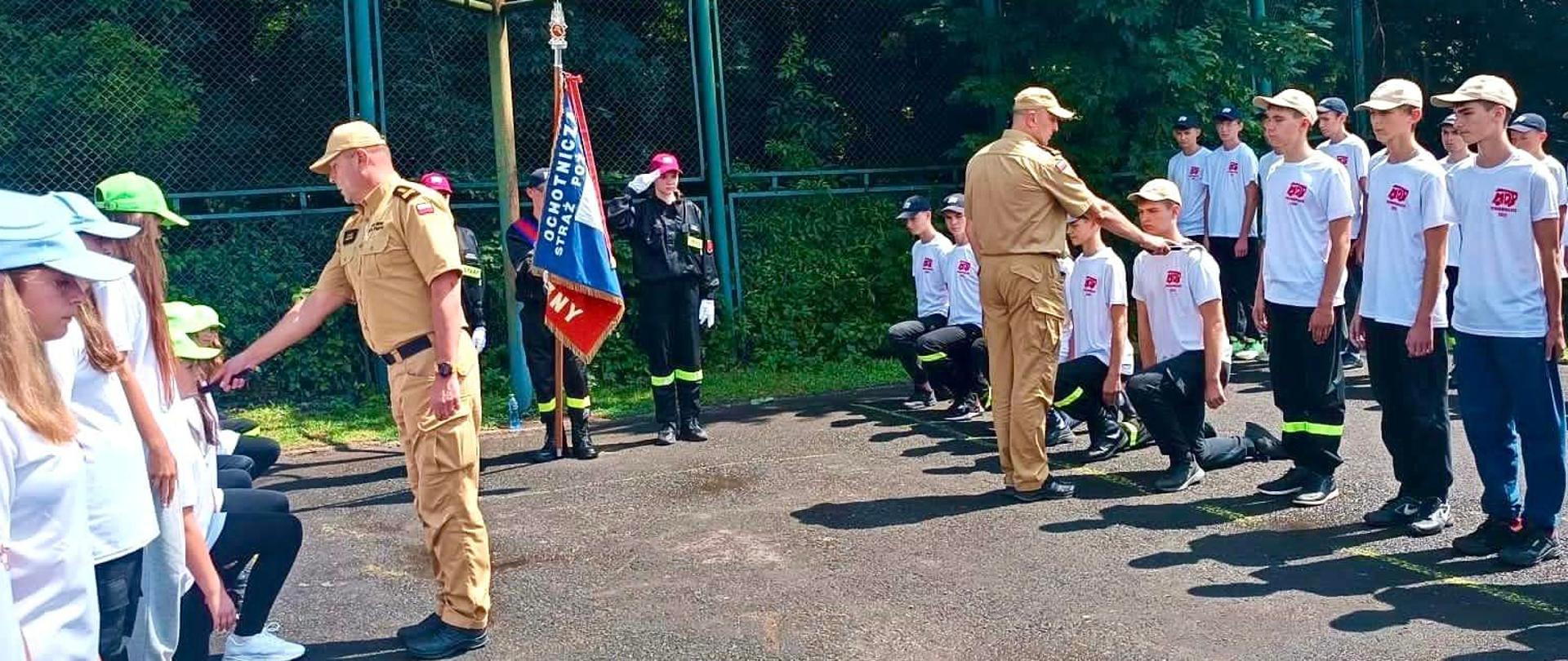 Zdjęcie zrobione na zewnątrz, na boisku sportowym, podczas uroczystego apelu kończącego turnus zgrupowania młodzieżowych drużyn pożarniczych. Dwóch oficerów Państwowej Straży Pożarnej pasuje przyjmuje ślubowanie młodych adeptów sztuki Pożarniczej. 
