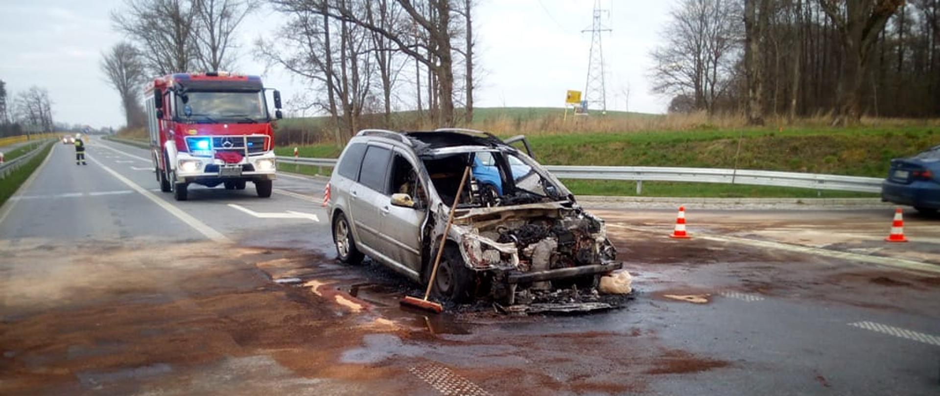 Samochód osobowy na drodze po pożarze w przedziale silnikowym pojazdu. Dokoła pojazdu rozsypany środek, który zneutralizowali rozlewisko substancji ropopochodnej. W tle widoczny samochód ratowniczo - gaśniczy.