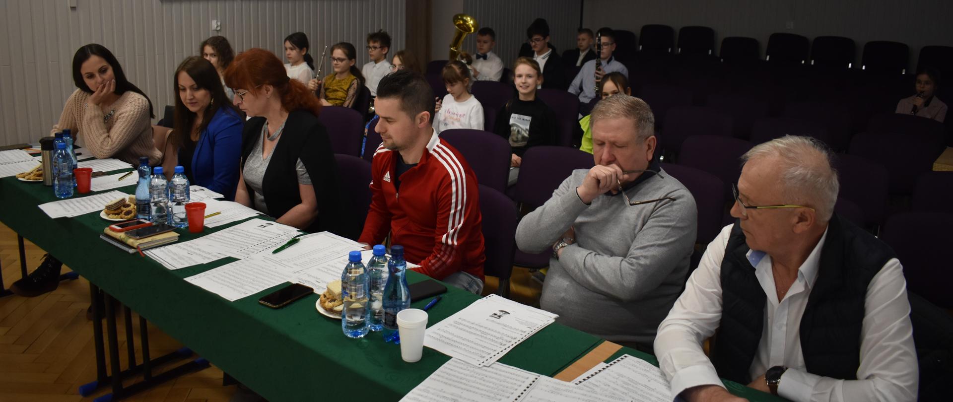 Na pierwszym planie przy zielonym stole siedzą nauczyciele szkoły oceniający uczniów trzy kobiety i trzech mężczyzn w różnokolorowych ubraniach. Za nimi w tle siedzą dzieci z instrumentami dętymi, na szarej ścianie zawieszony jest kolorowy obraz. 