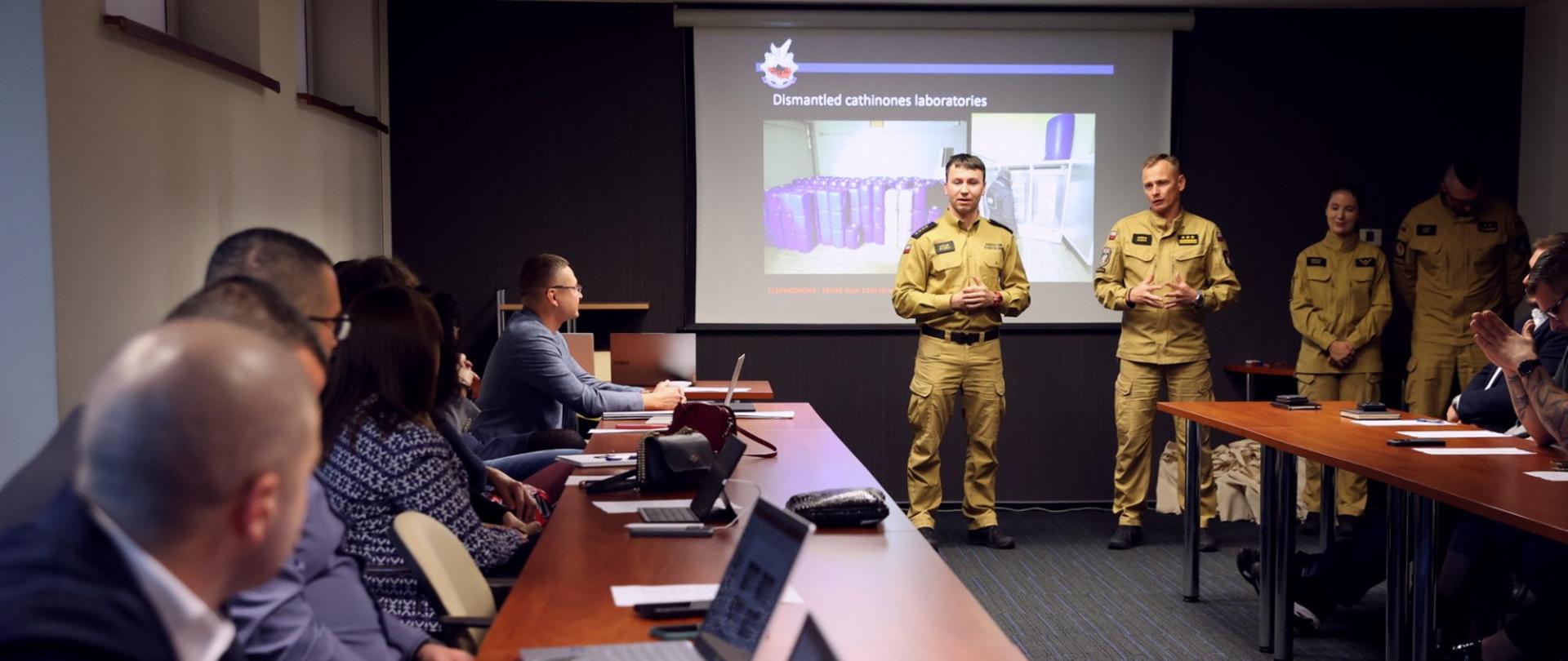 Sala konferencyjna, po lewej uczestnicy szkolenia siedzący za stołami podczas zajęć, w głębi stojący funkcjonariusze CS PSP. Za nimi ekran multimedialny z wyświetlonym slajdem prezentacji