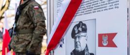 Po prawej stronie zdjęcia widać tablicę z wizerunkiem gen. broni Waldemara Skrzypczaka oraz informację pisemną o życiorysie generała. Tablica jest przepasana biało-czerwoną szarfą. Po lewej stronie zdjęcia widać sylwetkę żołnierza z bronią. W tle dwie flagi biało-czerwone i drzewa.