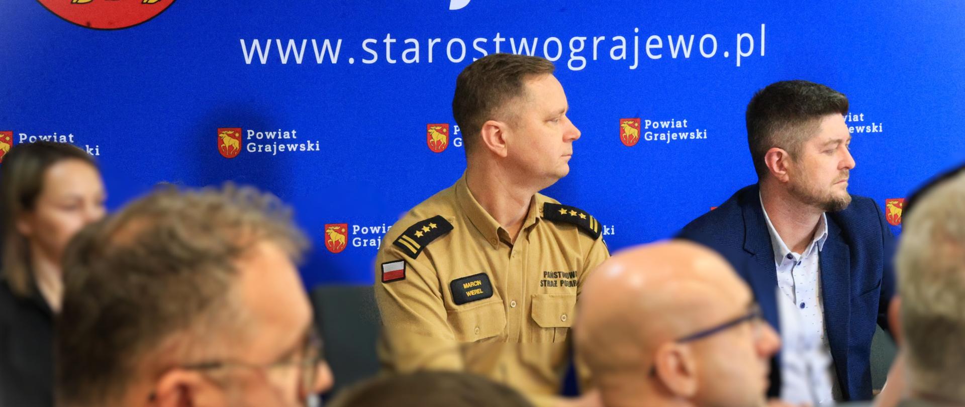 Grajewska konferencja dot. realizacji Programu Ochrony Ludności i Obrony Cywilnej - stół prezydialny przy którym siedzi zastępca podlaskiego komendanta wojewódzkiego PSP, dyrektor WCZK PUW, w tle baner starostwa powiatowego w Grajewie