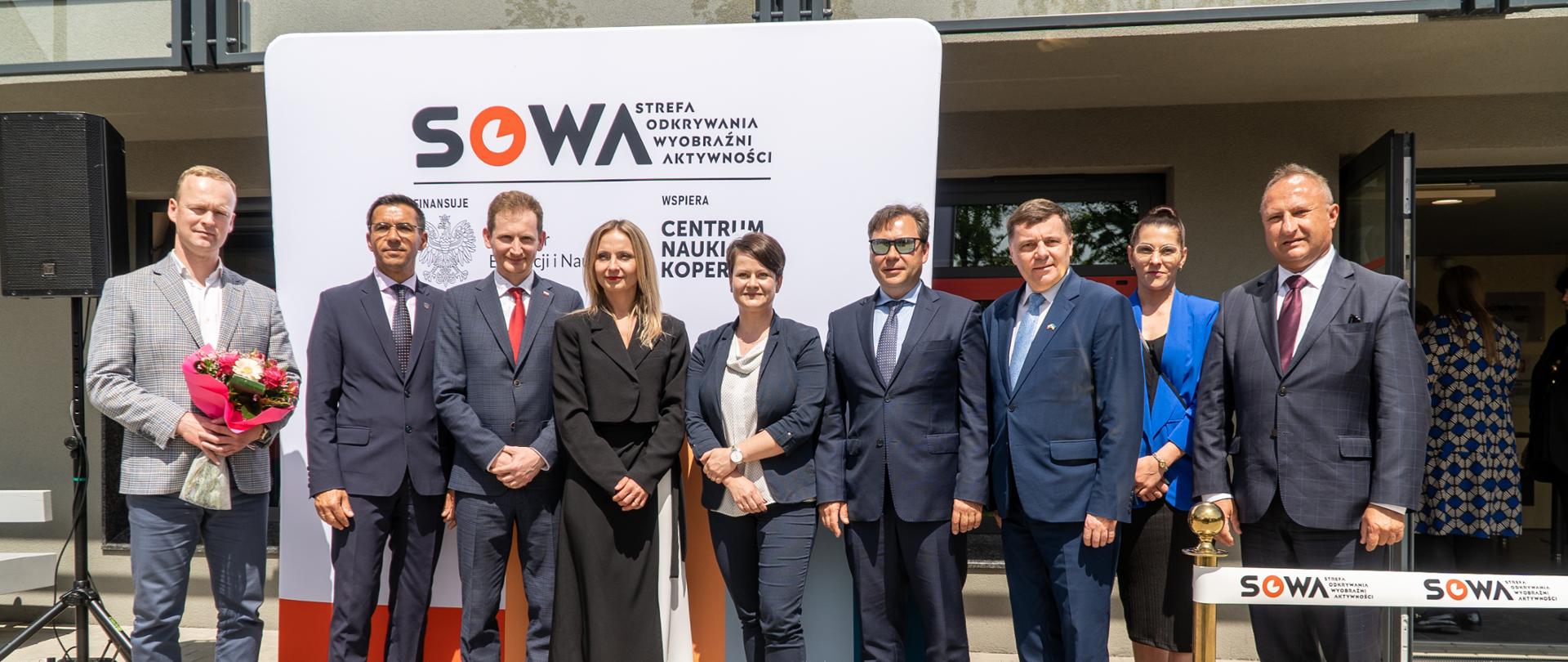 Na dworze w słońcu stoi w szeregu grupa elegancko ubranych osób, mężczyzna po lewej trzyma bukiet kwiatów, za nimi ścianka z napisem SOWA, dalej wejście do budynku.