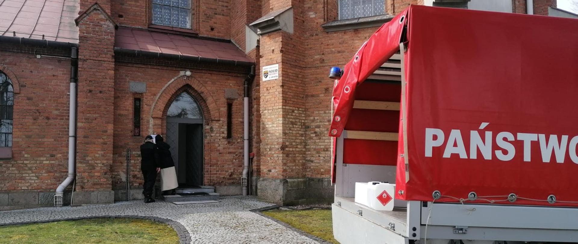 Samochód strażacki stoi zaparkowany przed kościołem. Strażak daje do podpisania zakonnicy rozdzielnik. 
