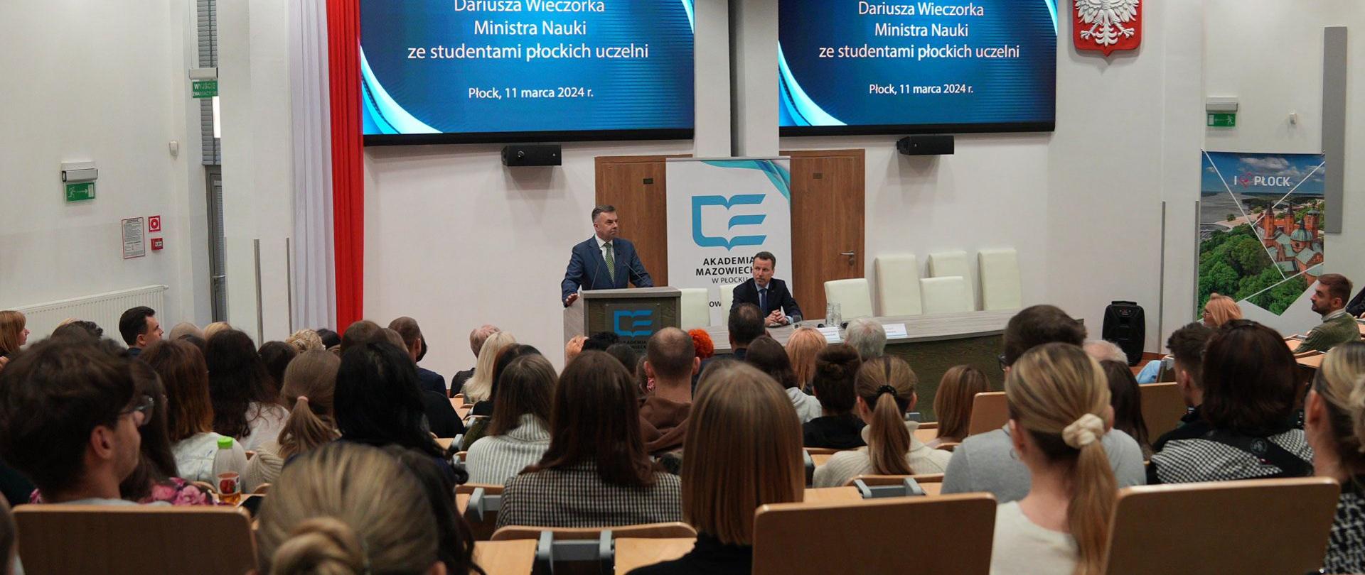 Widok od tyłu na salę wypełnioną siedzącymi w rzędach krzeseł ludźmi, z przodu za mównicą stoi minister Wieczorek i mówi do mikrofonu.