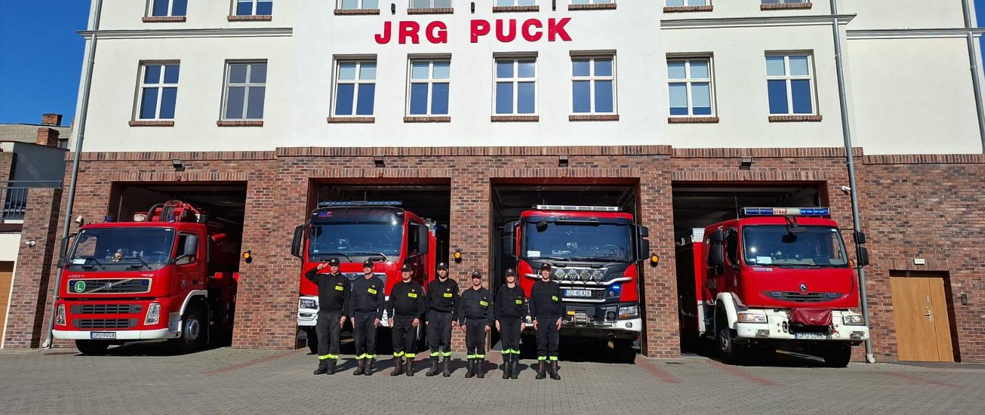 Funkcjonariusze z I zmiany pełniący służbę w dniu 1 sierpnia oddają honor uczestnikom Powstania Warszawskiego. Strażacy stoją w szeregu na baczność. Funkcjonariusze ubrani są w stroje koszarowe z czapkami na głowie. Za nimi ustawione są wozy bojowe będące na wyposażeniu Jednostki Ratowniczo Gaśniczej.