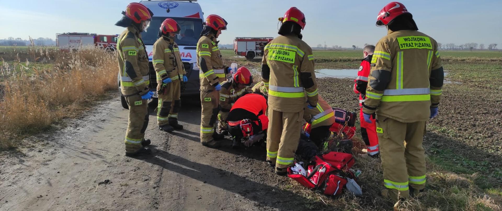 Na drodze utwardzonej stoi pięciu strażaków w mundurach i hełmach, obok ratownicy Zespołu ratownictwa Medycznego, pochylają sie nad osobą. Po bokach pole. W oddali pojazdy ratownicze.