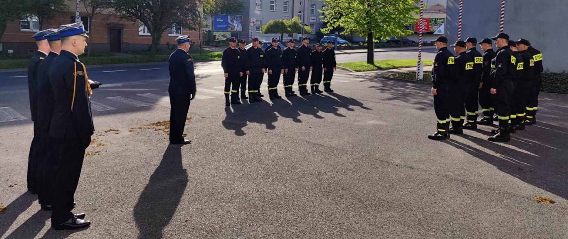 Zdjęcie przedstawia funkcjonariuszy Państwowej Straży Pożarnej. Funkcjonariusze ustawieni w dwóch szeregach zwróconych frontem ku sobie. Ubrani są w ubrania koszarowe w kolorze czarnym. Pomiędzy nimi stoi dowódca jednostki. Ubrany jest w galowy mundur. Strażacy ustawieni są w pozycji zasadniczej „na baczność”. Za dowódcą ustawiony jest dodatkowo poczet flagowy, który będzie dokonywał podniesienia flagi państwowej na masz. Poczet tworzy trzech funkcjonariuszy również ubranych w mundury galowe. Zdjęcie wykonane przed Jednostką Ratowniczo – Gaśniczą numer 1 w Radomiu. 