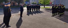 Zdjęcie przedstawia funkcjonariuszy Państwowej Straży Pożarnej. Funkcjonariusze ustawieni w dwóch szeregach zwróconych frontem ku sobie. Ubrani są w ubrania koszarowe w kolorze czarnym. Pomiędzy nimi stoi dowódca jednostki. Ubrany jest w galowy mundur. Strażacy ustawieni są w pozycji zasadniczej „na baczność”. Za dowódcą ustawiony jest dodatkowo poczet flagowy, który będzie dokonywał podniesienia flagi państwowej na masz. Poczet tworzy trzech funkcjonariuszy również ubranych w mundury galowe. Zdjęcie wykonane przed Jednostką Ratowniczo – Gaśniczą numer 1 w Radomiu. 