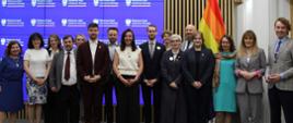 Na tle wbudowanego w ścianę z cienkich pionowych listewek wielkiego ekranu z napisem Ministerstwo Nauki i Szkolnictwa Wyższego stoi grupa kilkunastu osób, pośrodku wiceminister Zioło-Pużuk, za nimi z prawej tęczowa flaga.