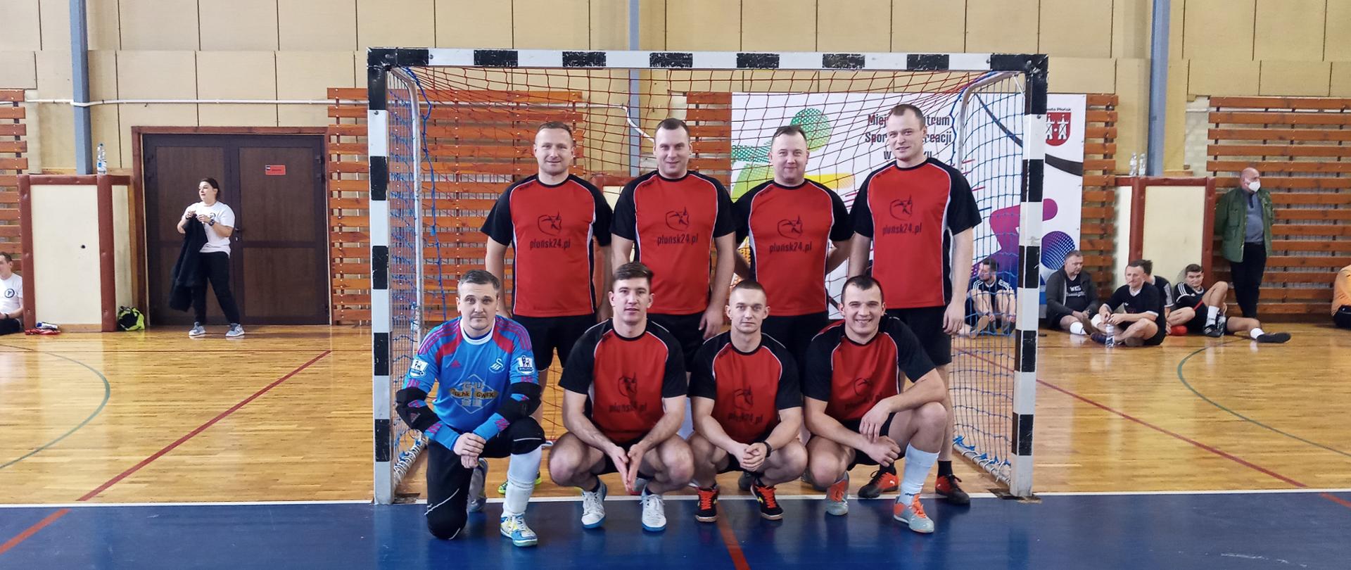 Na tle brami stoi 8 zawodników piłkarskich w strojach sportowych. W pierwszym rzędzie kuca czterech zawodników, w drugim rzędzie stoi czterech zawodników. W tle bramka do piłki ręcznej.