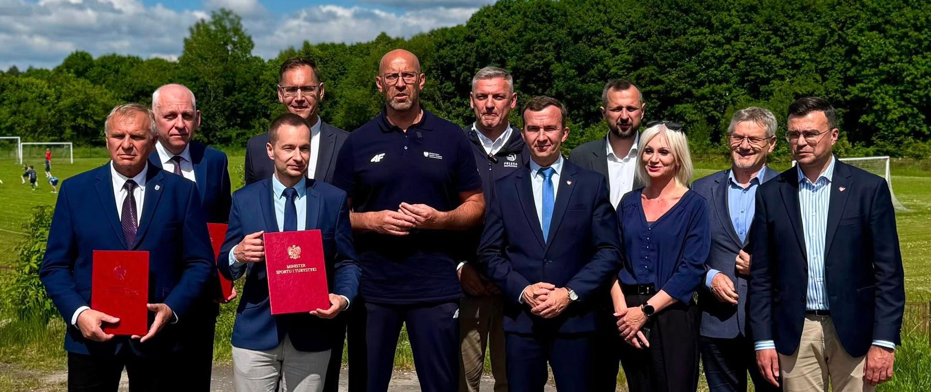 grupa oficjeli ubrana w stonowane kolory pozuje do zdjęcia trzymając czerwone teczki z orłem 
